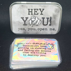 May include: A silver tin with the words "HEY YOU! yes, you. open me." printed on it. The tin contains a stack of cards with a rainbow swirl background and the text "you are amazing because you keep getting up, showing up, and facing the demons that are trying to destroy you."