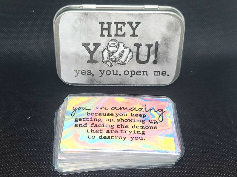 May include: A silver tin with the words "HEY YOU! yes, you. open me." printed on it. The tin contains a stack of cards with a rainbow swirl background and the text "you are amazing because you keep getting up, showing up, and facing the demons that are trying to destroy you."