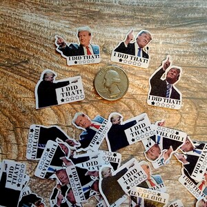 K&ouml;nnte beinhalten: Eine Sammlung von Aufklebern mit einem Mann im Anzug mit dem Text "I DID THAT!" und einer Sternebewertung. Ein US-Vierteldollar ist ebenfalls vorhanden. Die Aufkleber sind auf einer Holzoberfl&auml;che verstreut.