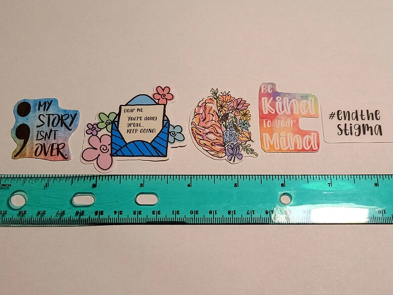 May include: Four colorful stickers with positive messages. The first sticker is a watercolor design with a semicolon and the text "My story isn't over". The second sticker is a blue envelope with a white heart and the text "Dear me, you're doing great... keep going!". The third sticker is a brain with flowers and the text "Be kind to your mind". The fourth sticker is a white sticker with the text "#endthe stigma".