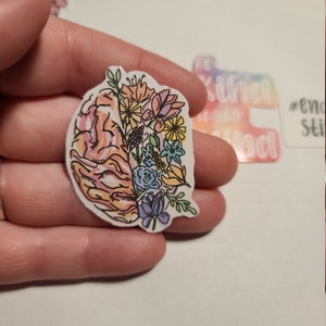 May include: A sticker featuring a colorful floral design inside a pink brain outline. The sticker is on a white background.