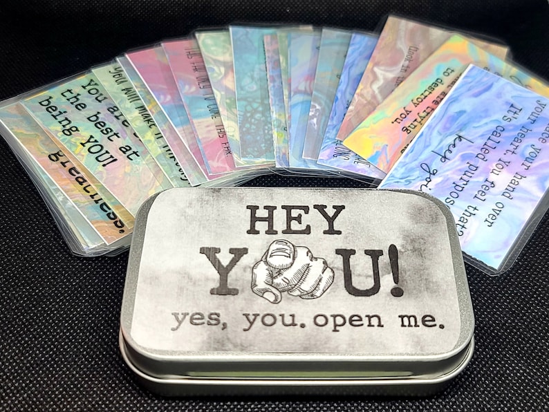 May include: A silver tin with the words "HEY YOU! yes, you. open me." printed on it. The tin contains a set of colorful cards with inspirational messages.