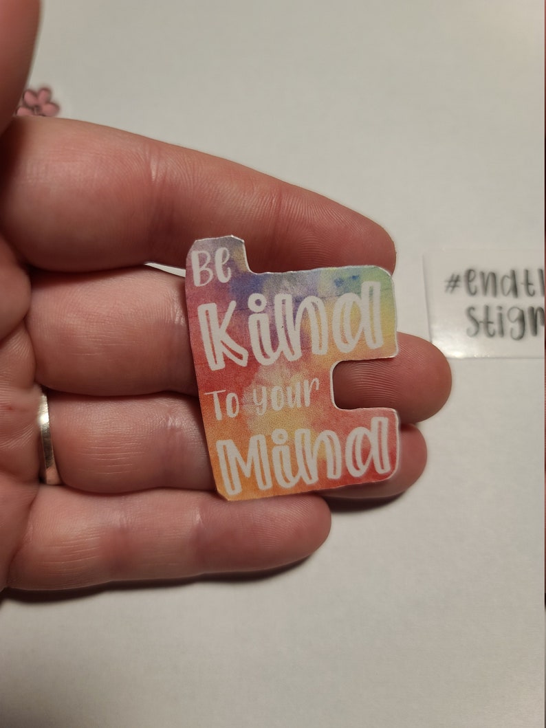 May include: A colorful sticker with the text "Be Kind To Your Mind" in white lettering on a rainbow watercolor background. The sticker is in the shape of a rectangle with rounded corners. The hashtag "#endthestigma" is visible in the background.