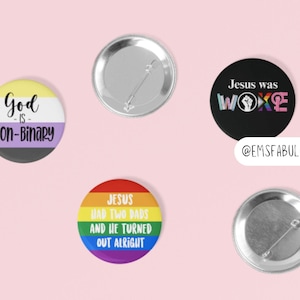 May include: Set of six 2.25 inch button pins with various LGBTQ+ themed messages. The pins feature a rainbow, a gender neutral flag, and a black background with white text. The text on the pins reads "God is non-binary", "Jesus had two dads and he turned out alright", and "Jesus was woke".