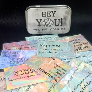 May include: A silver tin with the words "HEY YOU! yes, you. open me." printed on it. The tin contains a set of colorful affirmation cards with inspirational messages such as "Happiness. It's not a chance. It's a choice." and "You are a warrior for everything you overcome."