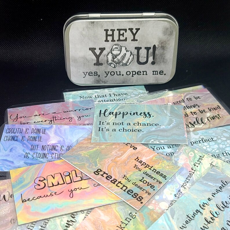 May include: A silver tin with the words "HEY YOU! yes, you. open me." printed on it. The tin contains a set of colorful affirmation cards with inspirational messages such as "Happiness. It's not a chance. It's a choice." and "You are a warrior for everything you overcome."