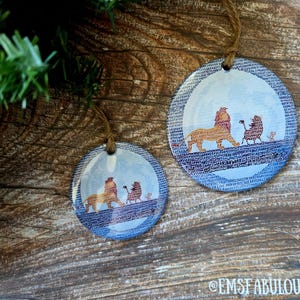 May include: Two round ornaments with a Lion King theme. The ornaments feature a lion, a baboon, and a small lion cub walking on a bridge. The background is a blue sky with a border of text. The ornaments hang from twine.