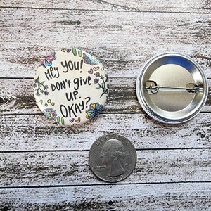 May include: A white button with a colorful floral design and the text "Hey you! Don't give up. Okay?" The back of the button is silver with a pin.