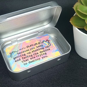 May include: A silver metal tin with a colorful card inside. The card has a motivational quote that reads: "you are amazing because you keep getting up, showing up, and facing the demons that are trying to destroy you."