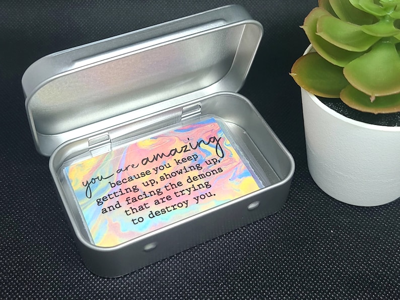 May include: A silver metal tin with a colorful card inside. The card has a motivational quote that reads: "you are amazing because you keep getting up, showing up, and facing the demons that are trying to destroy you."