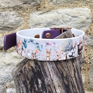 Könnte beinhalten: Ein weißes Hundehalsband mit einem verspielten Aufdruck von Waldtieren, Luftballons und Partyhüten. Das Halsband hat einen lila Lederakzent und kupferfarbene Beschläge. Das Halsband liegt auf einem verwitterten Baumstumpf.