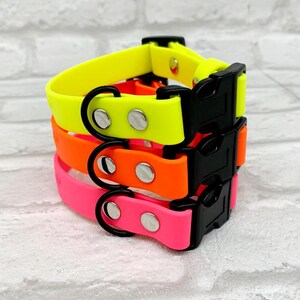 K&ouml;nnte beinhalten: Ein Stapel aus drei wasserfesten Halsb&auml;ndern in Neongelb, leuchtendem Orange und Pink. Jedes Halsband verf&uuml;gt &uuml;ber eine schwarze Kunststoff-Steckschnalle, einen schwarzen D-Ring aus Metall und zwei silberfarbene Nieten zur Befestigung.