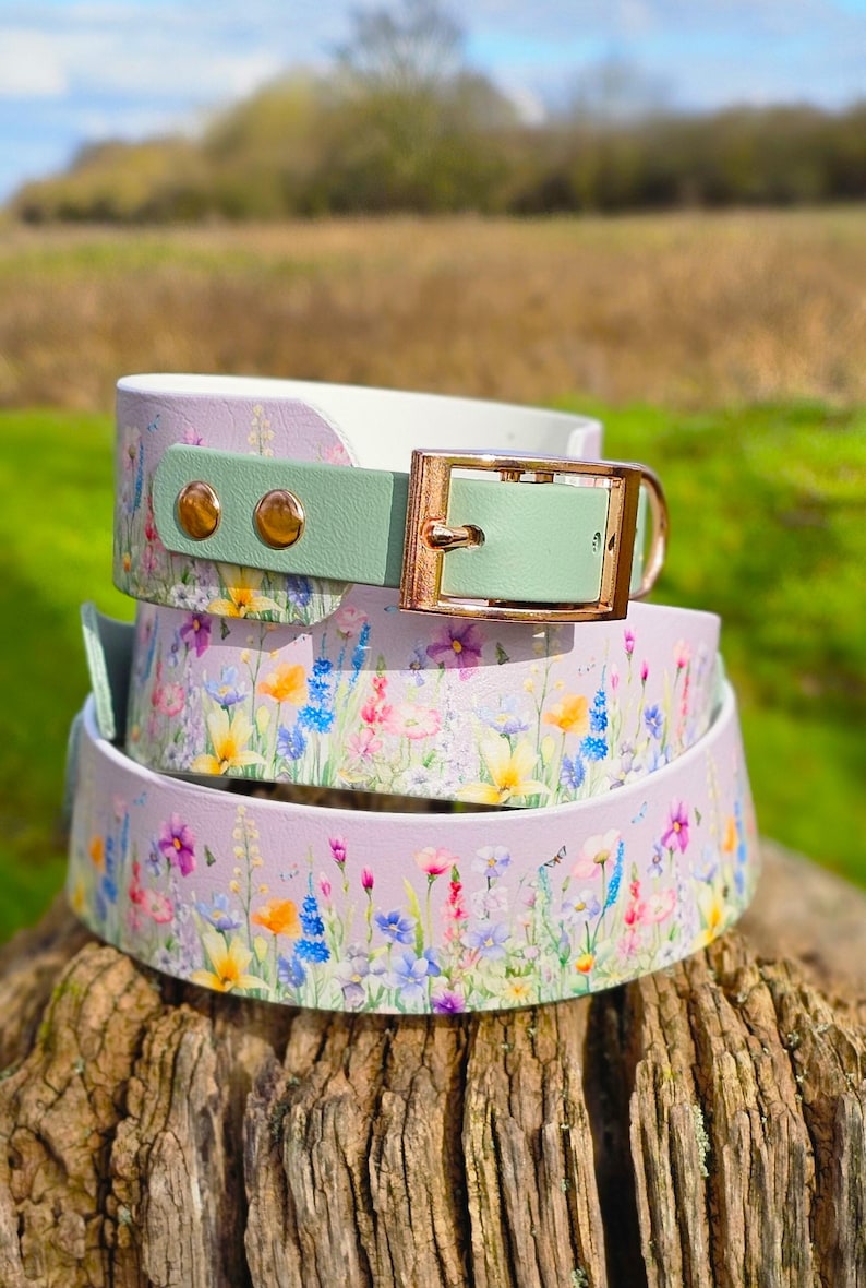 May include: Three dog collars stacked on a wooden surface. The collars feature a floral pattern with purple, yellow, and blue flowers. Each collar has a light green buckle and gold-colored hardware.