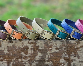 May include: A row of eight waterproof collars in black, brown, orange, olive, beige, teal, blue, and pink. Each collar features a brass-toned metal buckle and D-ring, with visible water droplets on the surface of the material.