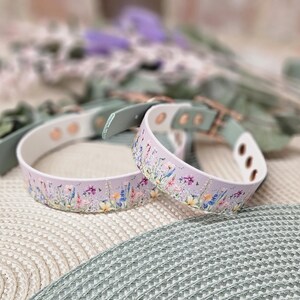 May include: Two lavender dog collars with a floral pattern. The collars have a white interior and are accented with green leather and gold-colored hardware. The collars are resting on a textured surface with blurred greenery in the background.
