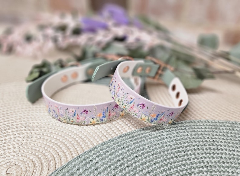 May include: Two lavender dog collars with a floral pattern. The collars have a white interior and are accented with green leather and gold-colored hardware. The collars are resting on a textured surface with blurred greenery in the background.