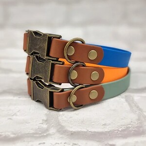 May include: Three stacked collars in blue, orange, and sage green. Each collar features a brown strap section, a brass-toned metal side-release buckle, a matching D-ring, and two brass-toned rivets securing the hardware to the colored strap.
