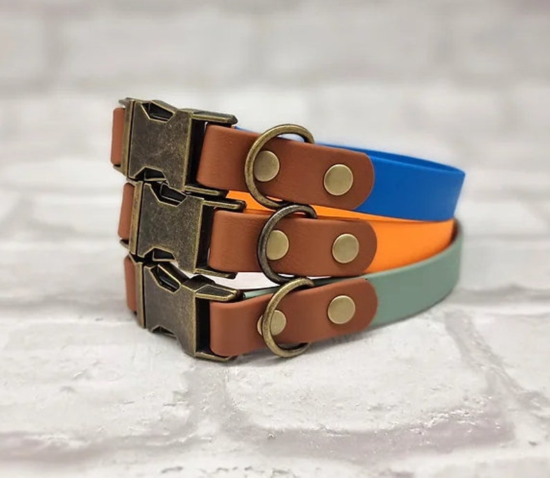 May include: Three stacked collars in blue, orange, and sage green. Each collar features a brown strap section, a brass-toned metal side-release buckle, a matching D-ring, and two brass-toned rivets securing the hardware to the colored strap.
