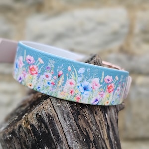Könnte beinhalten: Ein hellblaues Hundehalsband mit einem Blumendesign mit rosa, roten, lila und blauen Blumen. Das Halsband liegt auf einem Stück verwittertem Holz. Das Halsband hat eine weiße Innenseite und einen roségoldenen Verschluss.