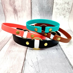May include: Three collars in black, coral, and turquoise, each featuring a central metal ring connected by two brass-coloured rivets. The bands are smooth and solid-coloured, with the black collar in the foreground, the coral collar behind it, and the turquoise collar positioned at the top.