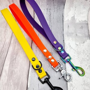 May include: Three short pet leashes in bright yellow, orange, and purple. Each leash features a loop handle, two metal rivets, a small D-ring, and a swivel snap hook. The yellow leash has black hardware, the orange has silver, and the purple has iridescent hardware.