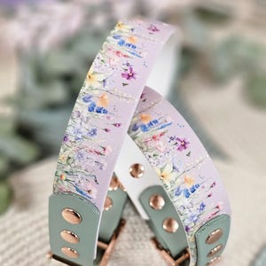 May include: Two floral-patterned dog collars. The collars feature a lavender background with a watercolor floral design in shades of pink, blue, yellow, and purple. The ends are sage green with rose gold hardware and white backing.