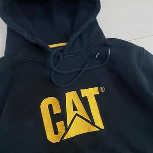May include: Navy blue hooded sweatshirt with the CAT logo in yellow. The sweatshirt has a drawstring hood and a kangaroo pocket. The CAT logo is embroidered on the front of the sweatshirt.
