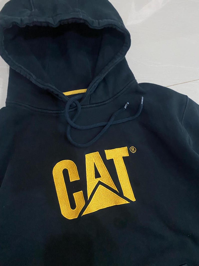 May include: Navy blue hooded sweatshirt with the CAT logo in yellow. The sweatshirt has a drawstring hood and a kangaroo pocket. The CAT logo is embroidered on the front of the sweatshirt.