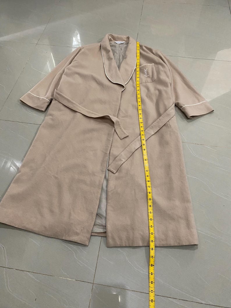 K&ouml;nnte beinhalten: Beigefarbener Bademantel mit wei&szlig;er Paspelierung und passendem G&uuml;rtel. Der Bademantel hat eine Tasche mit gesticktem Logo. Die L&auml;nge des Bademantels betr&auml;gt ca. 132 cm.