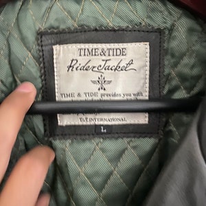 May include: Close-up of a black leather jacket's inner lining. The label reads "TIME&TIDE Rider Jacket" and "TIME & TIDE provides you with... T&T INTERNATIONAL". The lining is quilted in a diamond pattern and is green.