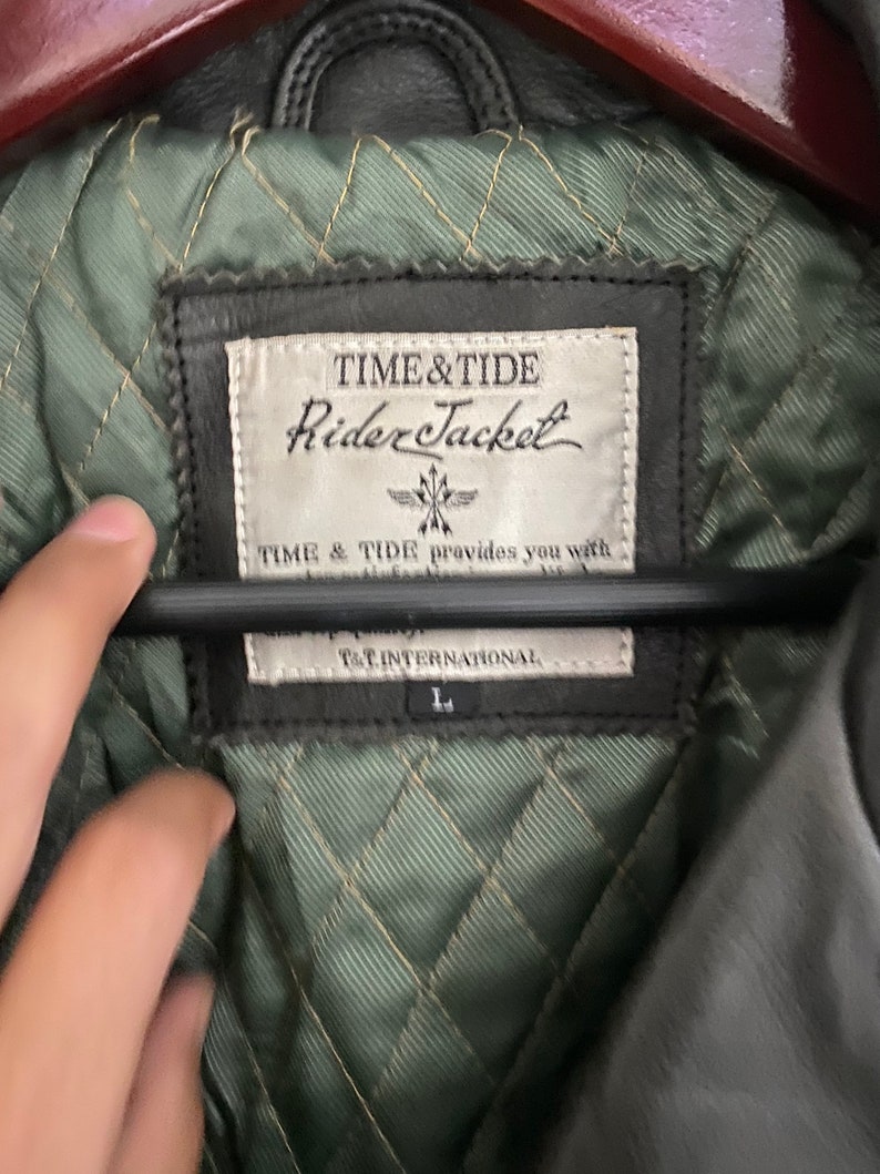 May include: Close-up of a black leather jacket's inner lining. The label reads "TIME&TIDE Rider Jacket" and "TIME & TIDE provides you with... T&T INTERNATIONAL". The lining is quilted in a diamond pattern and is green.