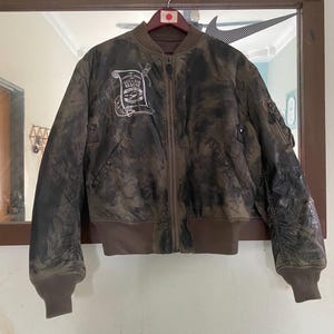 May include: A brown bomber jacket with a distressed, mottled appearance. The jacket features a ribbed collar, cuffs, and hem. A patch on the chest reads "SEA AIR RESCUE". The jacket is hanging on a wooden hanger.