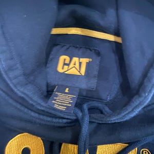 May include: Navy blue hooded sweatshirt with the CAT logo in yellow. The sweatshirt has a drawstring and a tag with the size and material information. The CAT logo is also embroidered in yellow on the front of the sweatshirt.