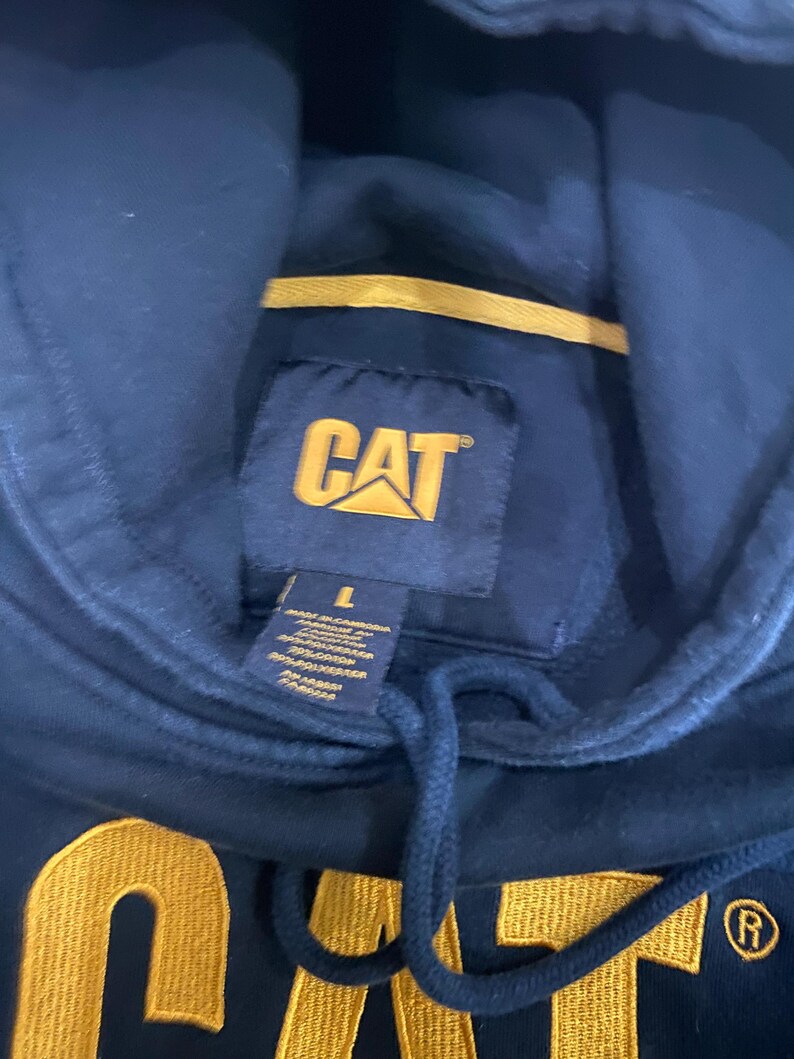 May include: Navy blue hooded sweatshirt with the CAT logo in yellow. The sweatshirt has a drawstring and a tag with the size and material information. The CAT logo is also embroidered in yellow on the front of the sweatshirt.