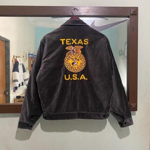Op de afbeelding: Een donkergrijze jas met de woorden "TEXAS" en "U.S.A." in gele letters. De achterkant van de jas heeft een cirkelvormig embleem met een gedetailleerd ontwerp. De jas hangt aan een hanger.