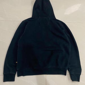 May include: A black hooded sweatshirt with a drawstring hood. The sweatshirt has long sleeves and a ribbed hem and cuffs. The back of the sweatshirt is plain, with a small square patch near the hood. The fabric appears to be a soft, medium-weight material.