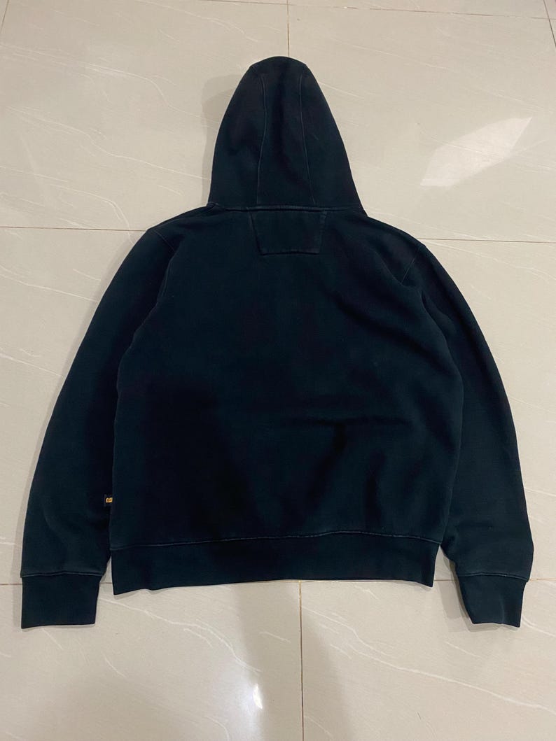 May include: A black hooded sweatshirt with a drawstring hood. The sweatshirt has long sleeves and a ribbed hem and cuffs. The back of the sweatshirt is plain, with a small square patch near the hood. The fabric appears to be a soft, medium-weight material.
