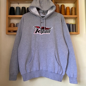 Pode incluir: Uma camisa de moletom cinza com capuz com um logotipo vermelho e preto do Ripon College na frente. O logotipo apresenta uma águia vermelha com uma asa preta e a palavra "Ripon" em preto.