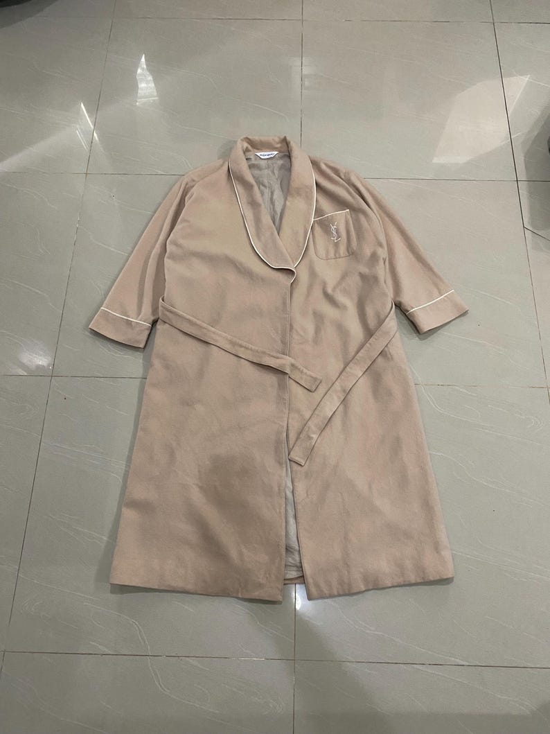 K&ouml;nnte beinhalten: Beiger Bademantel mit wei&szlig;er Paspelierung an Kragen, Manschetten und Tasche. Der Bademantel hat einen passenden G&uuml;rtel und ein kleines gesticktes Logo auf der Tasche. Der Bademantel ist aus weichem, leichtem Stoff.