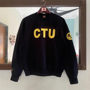 Op de afbeelding: Zwart sweatshirt met gele tekst "CTU" en een klein geel patch op de rechtermouw.