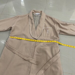 K&ouml;nnte beinhalten: Beiger Bademantel mit wei&szlig;er Paspelierung an Kragen und Revers. Er hat eine Tasche mit Logo auf der linken Brust und einen passenden G&uuml;rtel. Ein Ma&szlig;band liegt &uuml;ber dem Bademantel.