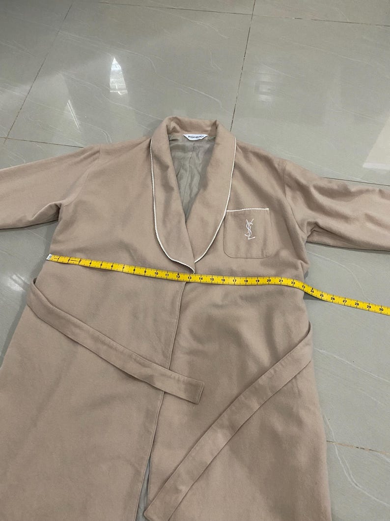 K&ouml;nnte beinhalten: Beiger Bademantel mit wei&szlig;er Paspelierung an Kragen und Revers. Er hat eine Tasche mit Logo auf der linken Brust und einen passenden G&uuml;rtel. Ein Ma&szlig;band liegt &uuml;ber dem Bademantel.