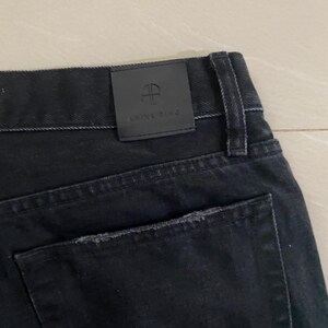 Puede incluir: Vaqueros de mezclilla negra con un bolsillo trasero desgastado y un parche de cuero negro con el texto "ANNE RING" y un logotipo.