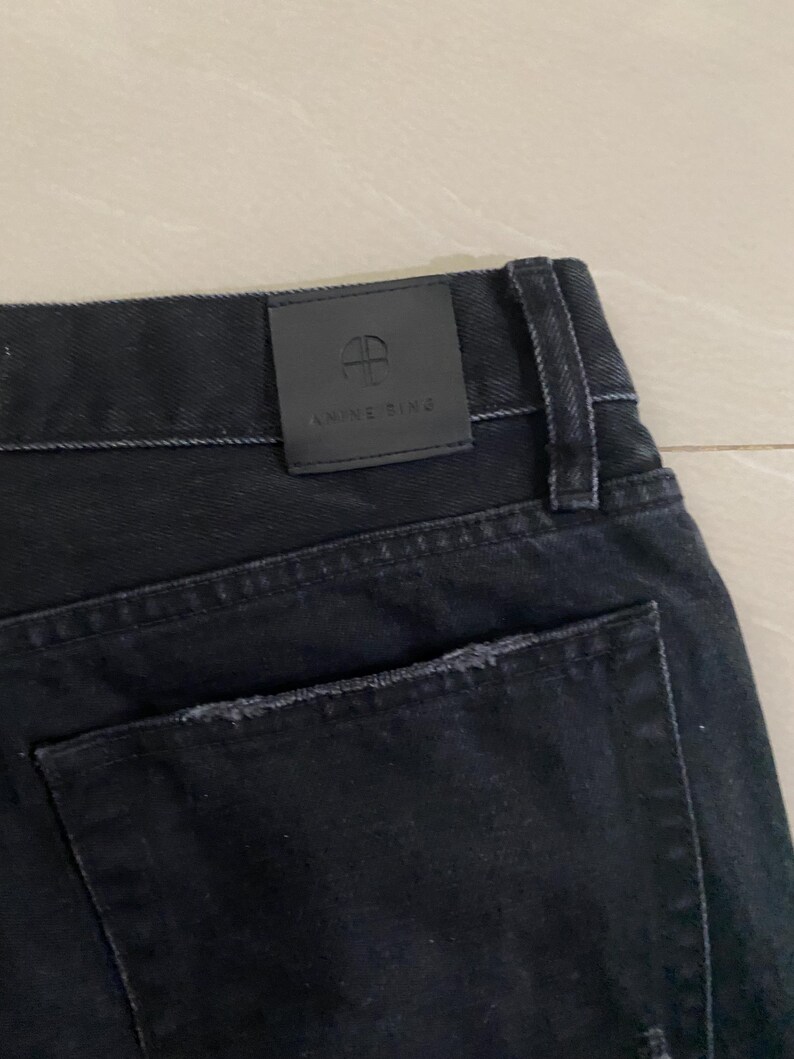 Puede incluir: Vaqueros de mezclilla negra con un bolsillo trasero desgastado y un parche de cuero negro con el texto "ANNE RING" y un logotipo.