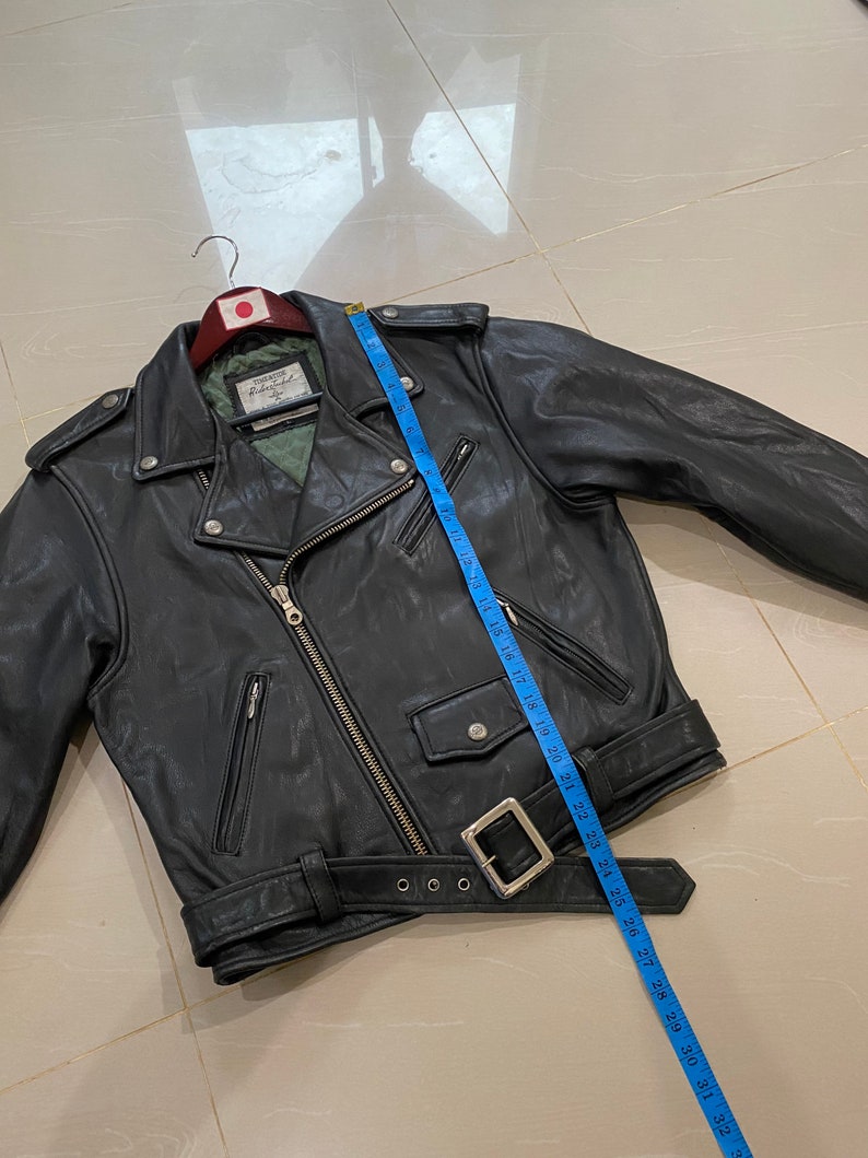 May include: Black leather motorcycle jacket with a zipper closure, two pockets, and a belt. The jacket is hanging on a hanger with a Japanese flag.