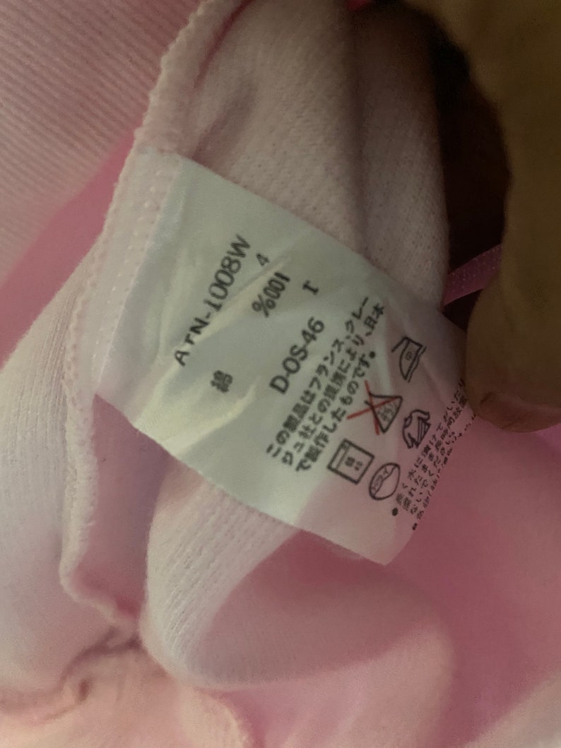 Puede incluir: Una etiqueta blanca con texto negro unida a una prenda rosa. La etiqueta dice "ATN-1008W", "Algod&oacute;n 4%", "D-OS-46", y "Este producto fue fabricado en Jap&oacute;n en colaboraci&oacute;n con la empresa francesa "Creations", y est&aacute; hecho con materiales provenientes de Francia. No lo deje en remojo en agua durante largos per&iacute;odos de tiempo."