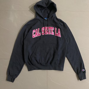 Könnte beinhalten: Ein dunkelgrauer Champion Hoodie mit einem pinkfarbenen "CAL STATE LA" Logo auf der Vorderseite. Der Hoodie hat eine Kapuze mit Kordelzug und ein kleines Champion Logo auf dem rechten Ärmel.