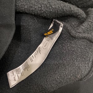 May include: Close-up of a black sweatshirt with a tag. The tag features the CAT logo in yellow and black, along with size information and fabric composition details. The sweatshirt is made of a cotton and polyester blend.