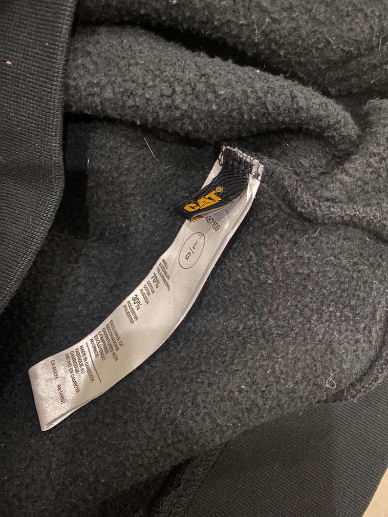 May include: Close-up of a black sweatshirt with a tag. The tag features the CAT logo in yellow and black, along with size information and fabric composition details. The sweatshirt is made of a cotton and polyester blend.