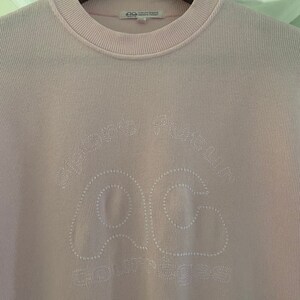 Puede incluir: Una camisa de manga larga de color rosa claro con un logotipo bordado en blanco que dice "Comme des Gar&ccedil;ons Play" y presenta un coraz&oacute;n con un contorno cosido en blanco.